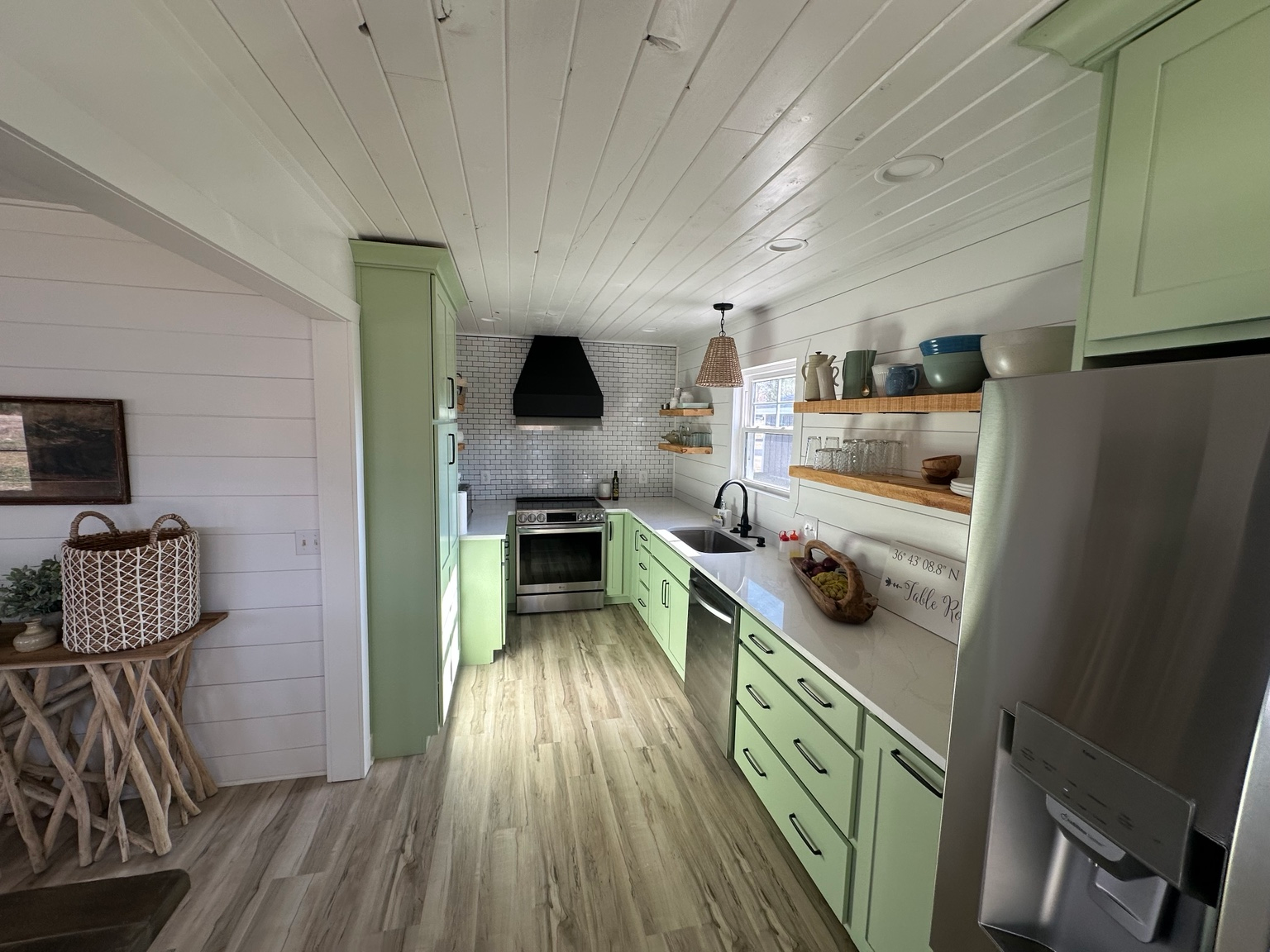 Kitchen with sage-green cabinetry, white countertops, open wood shelves, and a black range hood